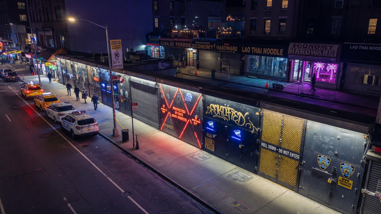 Aerial night view of protected NYC storefronts with illuminated custom steel doors creating a secure corridor on a busy commercial street, atmospheric lighting, cinematic 8K